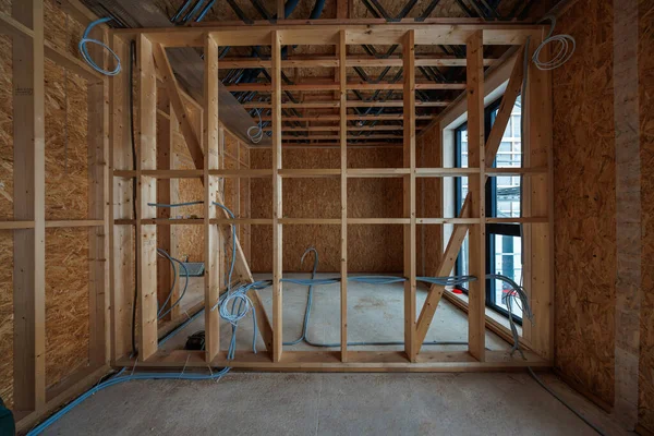 İç ahşap çerçeveli oda açıkta duran çiviler, OSB kılıfları, elektrik kabloları, çapraz destekleme, açık kirişlerdeki kanallar ve yumuşak doğal ışıkta uzun bir çift pencere gösteriyor..