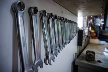 Krom cırcırtlak ve kombinasyon anahtarlar, dağınık bir bankın yanında boyutuna göre sıralanmış beyaz bir tahta üzerinde asılı, gündüz ışığı açık bir kapıdan girer ve yansıtır.