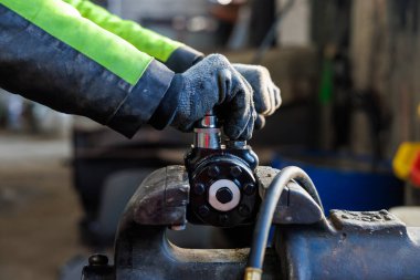 Yüksek görüşlü kolları olan bir teknisyen mengenede siyah bir vana sıkıyor ve yakında bir hortum kıvrılıyor. Yan ışık ve yüzeysel alan derinliği yağ ve metal dokularını ortaya çıkarıyor.