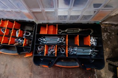 İki plastik kutu, yıpranmış bir atölye tezgahında, düzgün sıralanmış döner kollar, D prangaları, karabinalar, göz cıvataları, S kancaları, ve turuncu bölmelerde klipslerle açık durur..