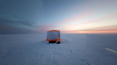 Turuncu ve beyaz bir sığınak ufukta donmuş bir gölde duruyor. Kameranın uzun gölgeli soğuk ışıkta yavaşça hareket ettiği kızak ve ayak izleri ona doğru gidiyor..