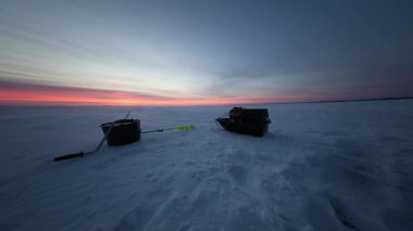 Donmuş bir gölün üzerinde tek başına bir buzda balık avı düzeneği var. Sarı bir çengelli, çubuklu siyah bir kova ve şafak öncesi ışık ve kırmızı turuncu ufukta desenli bir buzun üzerinde duran bir kızak..