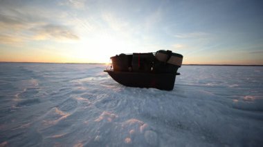 Alet kutuları ve soğutuculu plastik kızak kalın göl buzunun üzerinde duruyor. Sıcak altın ışık, uzun gölgeler ve geniş açı gündoğumunda ya da günbatımında balık avlamaya hazırlandığını gösteriyor..