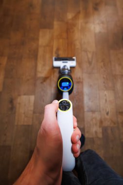 Bir el, elektrik ve oklarla ince beyaz bir uzaktan kumandayı tutarken kablosuz bir elektrikli süpürge, ahşap zemin üzerinde mavi bir LED ölçüm gösterir, dikey atış, sığ alan derinliği.