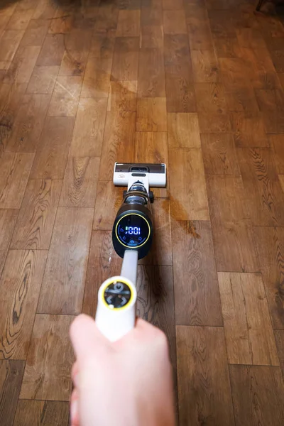 Biri beyaz kablosuz yer temizleyicisini orta boy ahşap bir döşeme üzerine itiyor. LED ekranda 100 yazıyor. Sarı aksanlar ve kuru bir çizgi kapalı alanda görülebilir..