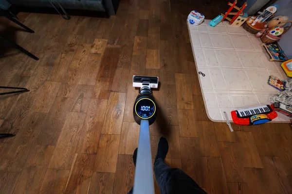 Bir insan kablosuz sopa süpürgesiyle yerleri süpürür. Oturma odasında oyun hasırı, oyuncak klavye ve tahta oyuncaklarla yüzde yüz gösterilir..