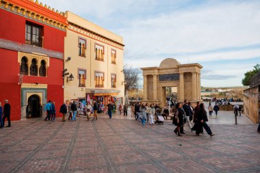 Gruplar tuğla plazadan geçerek üç kemerli kumtaşı geçidine, kemerli pencereli Fas tarzı cephelere ve sıcak öğleden sonra ışık saçan kaplamalı hediyelik eşya tezgahlarına doğru yürür..