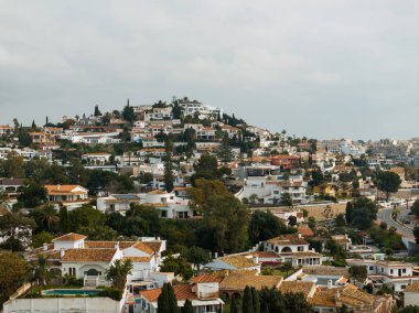 İspanya 'nın Marbella şehrinde terracotta çatısı olan beyaz boyalı villalar yuvarlak bir tepeye tırmanıyor. Palmiyeler, selvi ağaçları, havuzlar ve kıvrımlı bir kıyı yolu gün ortası bulutları altında görünür..