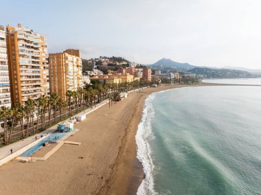 Malaga, İspanya 'daki La Malagueta Sahili kıvrımlı bir kıyı şeridi, palmiye güvertesi, sıcak deniz tarlası kuleleri, Gibralfaro Dağı, yumuşak erken aydınlık, nazik dalgalar, aylaklar ve bisikletçiler gösteriyor..