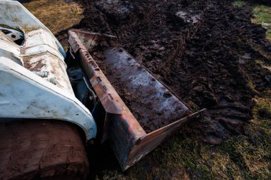 Paslı kovayla beyaz fren dümeni doymuş toprakta, derin lastik izlerinde ve raylarda çalkalanmış kil ve bıçakta, sıcak yan ışık, sıkı zemin seviyesinde hareket eder..