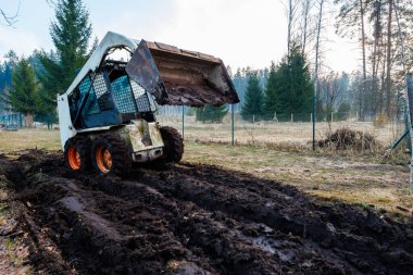 Paslı kovayla yükseltilmiş küçük bir kayma sığırı, çitlerle çevrili bir kırsal bölgedeki ıslak toprağı her zaman yeşil ağaçlarla paslandırır. Çamurlu tekerlekler ve taze izler serin gün ışığında aktif toprak hareketlerini gösteriyor..