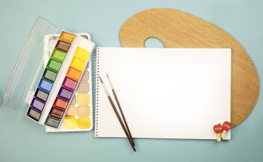 Okula dönelim. Mavi arka planda bir eskiz kitabı, mavi arkaplanda ahşap bir palet, mavi arkaplanda fırçalı suluboya resimler. Boya takımı. Öğrenme kavramı.