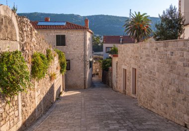 Rab, Hırvatistan - 26 Ağustos 2025: Eski taş binalar ve yeşilliklerle kaplı dar taşlarla kaplı bir cadde, açık mavi gökyüzü altındaki tepelerin uzak bir manzarasına yol açıyor.