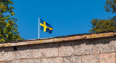 Vaxholm, İsveç - 13 Eylül 2025: Ağaçlı ve mavi gökyüzü olan taş bir duvarın üzerinde dalgalanan İsveç bayrağı.