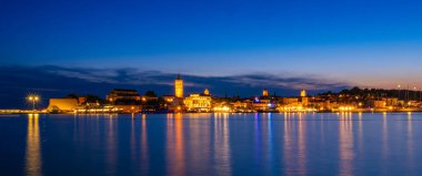 Rab, Hırvatistan - 25 Ağustos 2025: Derin mavi bir gökyüzünün altında sakin suları yansıtan aydınlık binaları olan sakin bir kıyı kasabasının gece manzarası.