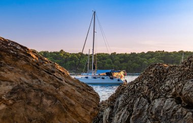 Rab, Hırvatistan - 25 Ağustos 2025: Ormanlı arka planda kayalık kıyılara demir atmış yelkenli.