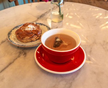 Kırmızı bir fincan çay, çay poşeti mermer bir masanın üstünde, pastanın yanında..