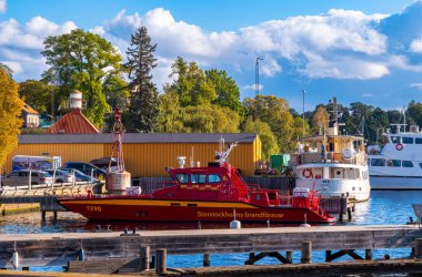 Stockholm, İsveç - 3 Ekim 2025: Kırmızı bir kurtarma botu arka planda sarı bir bina ve ağaçlarla limana yanaştı.