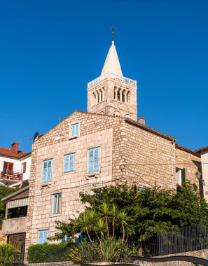 Rab, Hırvatistan - 26 Ağustos 2025: Mavi panjurlu taş bina ve açık gökyüzüne karşı bir kilise kulesi.