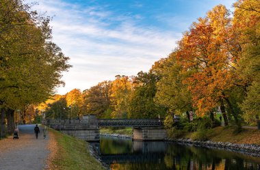 Stockholm, İsveç - 18 Ekim 2025: Renkli ağaçlar ve yürüyüş yolu ile çevrili sakin bir nehrin üzerinde köprüsü olan manzaralı sonbahar manzarası.