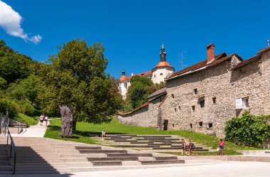 Skofja Loka, Slovenya - 30 Temmuz 2025: Taş binalar, bir kilise ve açık mavi gökyüzü altında basamakları ve ağaçları olan tarihi bir Avrupa kasabasının manzarası.