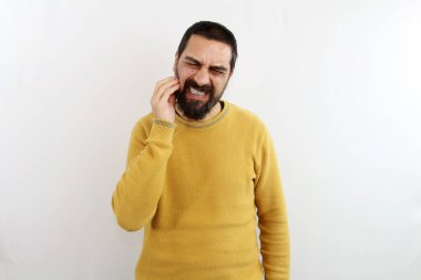 İzole edilmiş beyaz arka plandaki genç sakallı adam parmaklarıyla diş ağrısı için yanağını bastırıyor.