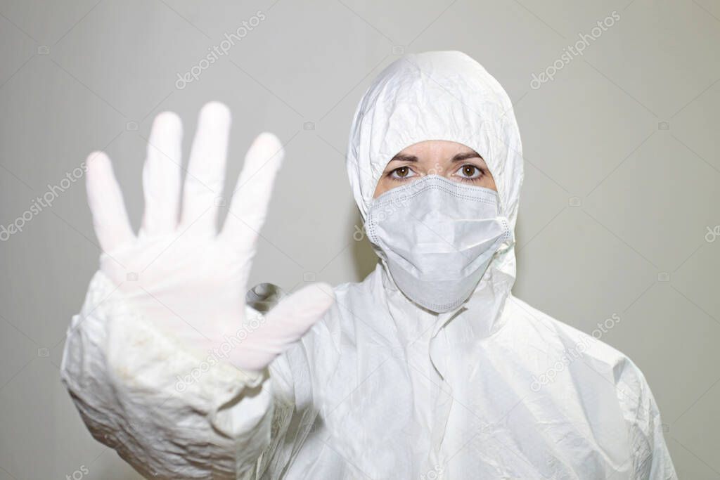 a female health worker with coverall and protective mask to fight coronavirus raises her hand as a sign of stopping. Public health concept