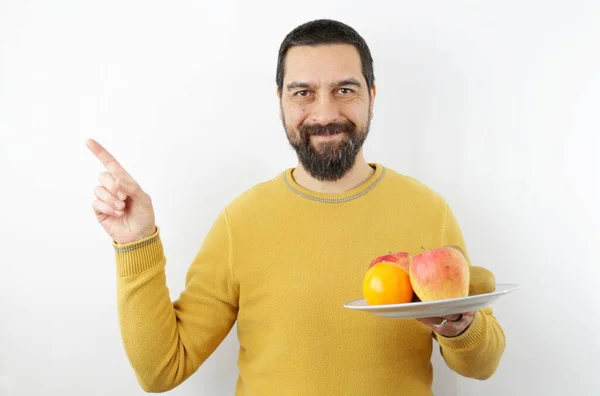 Beyaz arka planda gülümseyen sakallı bir adam bir tabak meyveyi parmağıyla kenarı gösteriyor.