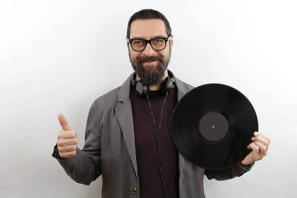 Sakallı ve gözlüklü adam elinde vinil disk ile gülümsüyor, mutlu ve pozitif, başparmağıyla mükemmel ve onay işareti yapıyor.