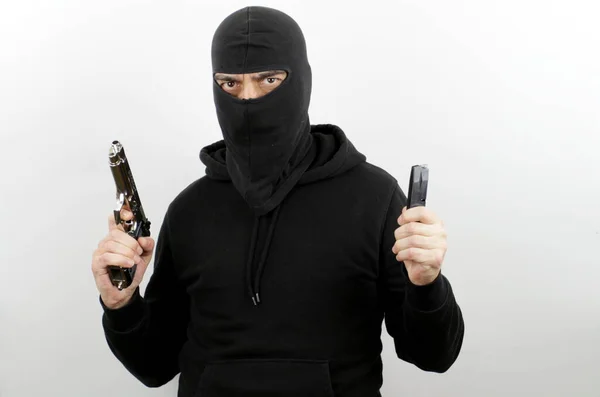 Criminal man wearing balaclava and hoodie holding pistol and magazine over isolated white background.
