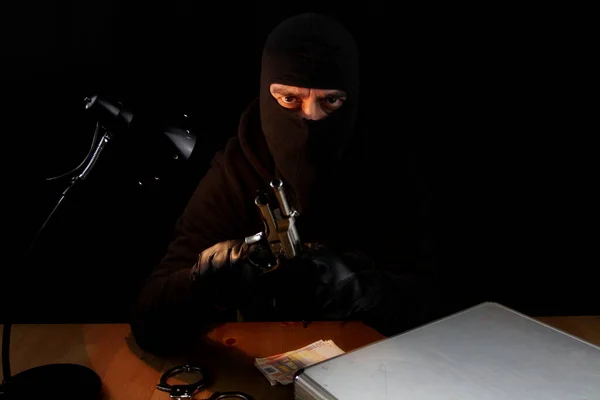 Man with balaclava holding gun. Sitting behind the desk looking at the camera, isolated on black background.