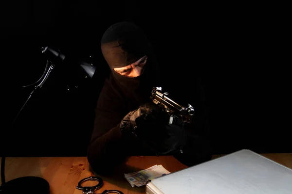 Man with balaclava holding gun. Sitting behind the desk poiting gun to the side, isolated on black background.