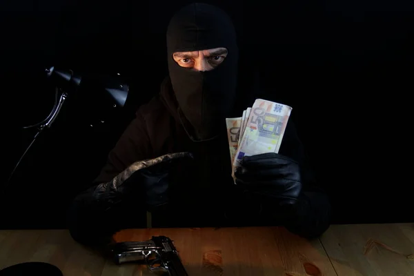 Contrabandist wearing on balaclava, holding Euro banknotes pointing with fingers. Sitting behind the desk, dealer, isolated on black background looking at the camera.
