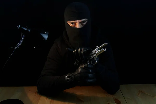 Criminal man with balaclava holding gun. Sitting behind the desk looking at the camera, isolated on black background.