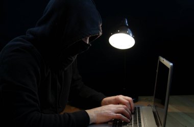 Kapüşonlu ve kar maskeli bir bilgisayar korsanı siyah arka planda izole bir şekilde dizüstü bilgisayarda oturuyor..