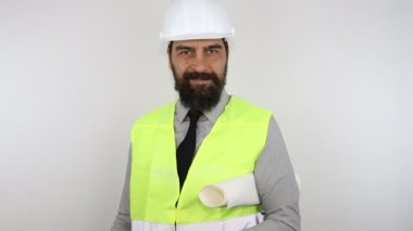 Mimar iş kaskı takıyor ve ceketi yansıtıyor. Kolunun altında taslak tutarak gülümsüyor ve elle sogn yapıyor..