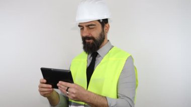 Sakallı mühendis iş kaskı takıyor ve ceketi yansıtıyor, tablet üzerinde tutuyor ve daktilo yazıyor..