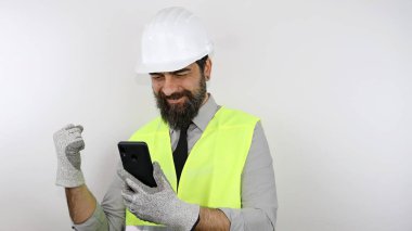 Sakallı mimar, kasketli ve yansıtıcı ceketli, Smatphone 'u tutan ve zaferde yumruk atan. Kazanan konsept