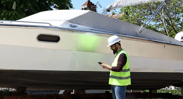 Foreman iş kaskı takıyor, test için tablet kullanıyor ve tersanedeki motorlu tekneyi kontrol ediyor. Çalışma konsepti.