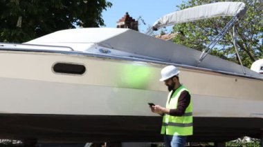 Foreman iş kaskı takıyor, test için tablet kullanıyor ve tersanedeki motorlu tekneyi kontrol ediyor. Çalışma konsepti.