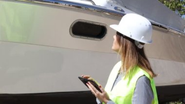 İş kaskı takan ve ceketi yansıtan kadın mühendis tersanedeki bir tekneyi test etmek ve kontrol etmek için tablet kullanıyor. Çalışma konsepti.