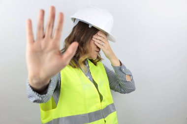 Kadın mimar ya da mühendis, kasket takıyor ve şok edici bir şekilde ceketini giyiyor. Yüzünü ve gözlerini mahcup bir ifadeyle örtüyor ve korku ifadesiyle dur hareketi yapıyor..