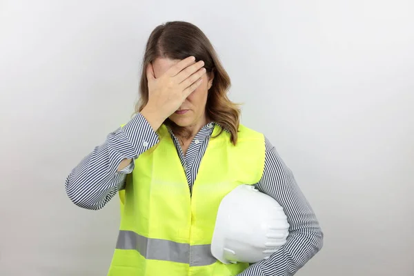 Female architect or engineer wearing reflective jacket holding safety helmet under arm, tired with hand on forehead feeling fatigue and headache. Stress and frustration concept.