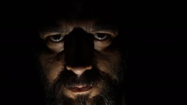 Karanlıktaki sakallı adam kameraya bakıyor. Karanlıkta hafif bir ışık.