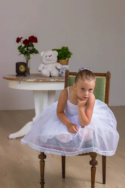 Cute little ballerina in a white dress sitting on wooden chair on a background with rose and bear — Stock Photo, Image