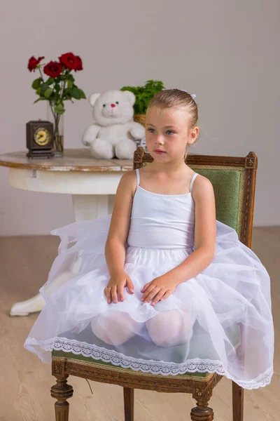 Young ballerina in a white dress sitting on wooden chair on a background with rose and bear — Stock Photo, Image