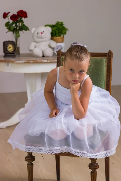 Cute little ballerina in a white dress sitting on wooden chair on a background with rose and bear — Stock Photo, Image