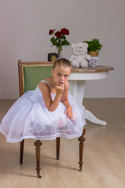 Niedliche kleine Ballerina in einem weißen Kleid sitzt auf einem Holzstuhl auf einem Hintergrund mit Rose und Bär — Stockfoto
