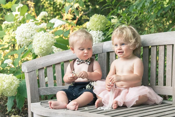 Junge und Mädchen in festlichem Kleid sitzen auf einer Holzbank in einem schönen Garten Stockbild