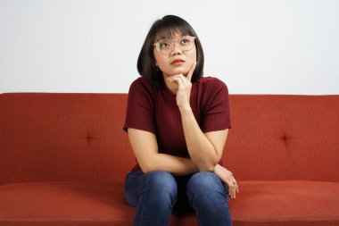 Kırmızı tişört ve mavi kot pantolon giyen güzel Asyalı kız kanepeye otur ve kafanı kaldır ve jestini düşün.